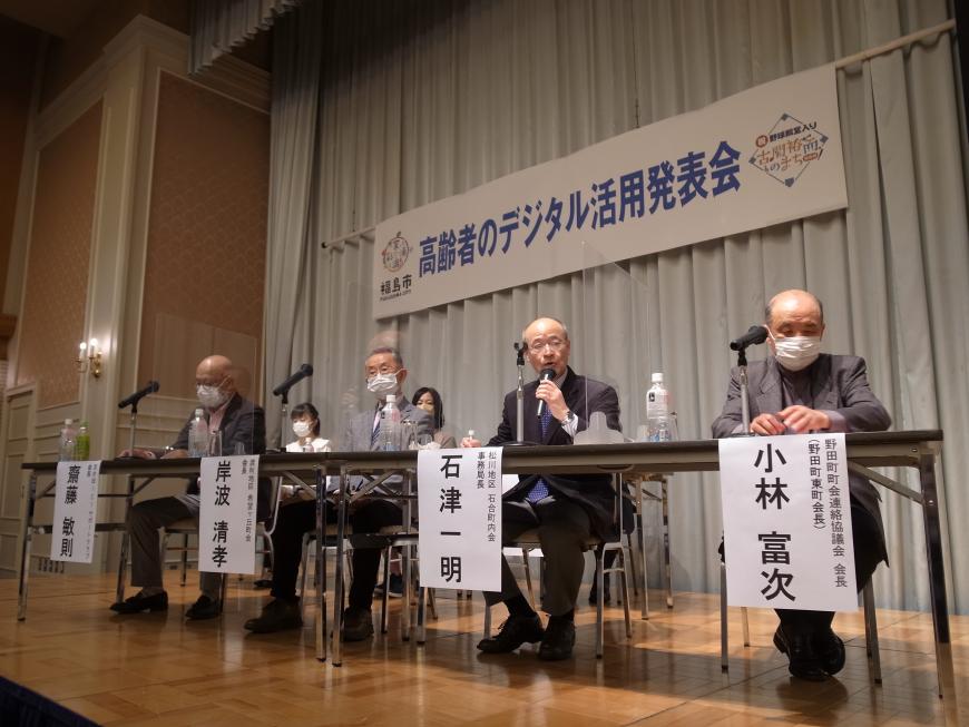 「高齢者のデジタル活用発表会」と書かれた横断幕が掲げられたステージに設置された長机の席に4名の男性が座り、右から二番目の男性が話をしている写真
