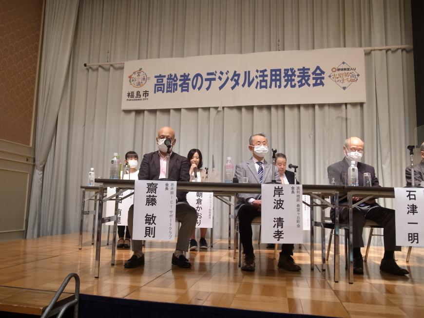 「高齢者のデジタル活用発表会」と書かれた横断幕が掲げられたステージの長机の席に座った3名の男性と、後列に3名の関係者が座っている写真
