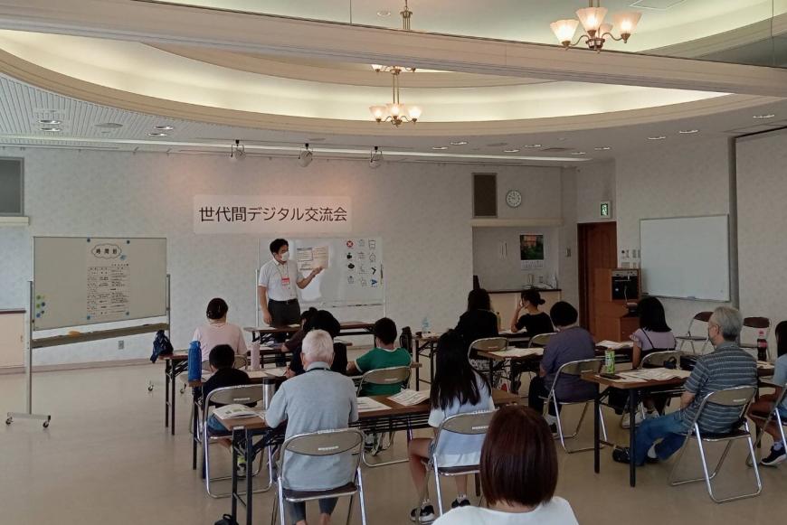 「世代間デジタル交流会」と書かれた室内前方に設置されたホワイトボードの前に立つ男性の話を、2人掛けの長机の椅子に参加者が座って聞いている様子の写真