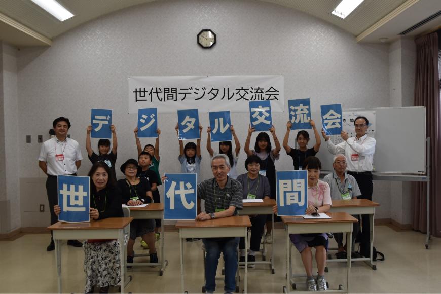 「世代間デジタル交流会」と書かれた幕が前方の壁に貼られた室内で、前列の席に座った方が一人一文字ずつ「世代間」の文字を持ち、後列に立つ子供たちと担当者の男性が「デジタル交流会」と書かれた文字を持っている写真