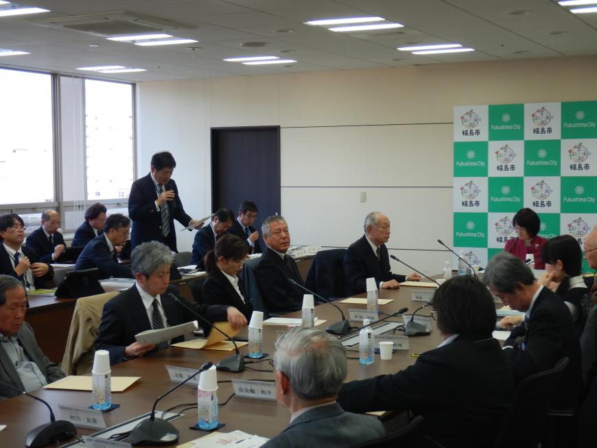 窓際に設置された机の前で1人の男性が資料を見ながら説明をしている写真