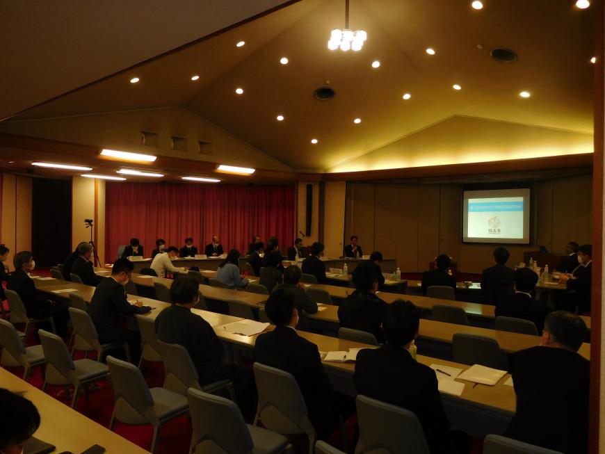 照明がおとされた室内で正面のモニターを見ながら会議に参加している人達を会場右斜め後方から写した写真