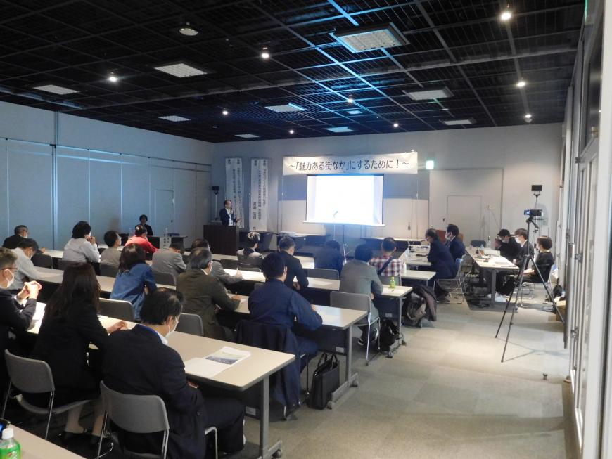 スクリーンの上に「魅力ある街なか」にするために!の幕が張られ演台に立つ渡邉氏が講演をしている様子を写した写真