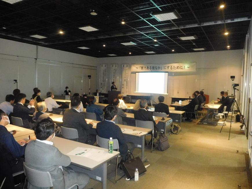 スクリーンの上に「魅力ある街なか」にするために!の幕が張られ演台に立つ石澤氏が講演をしている様子を写した写真