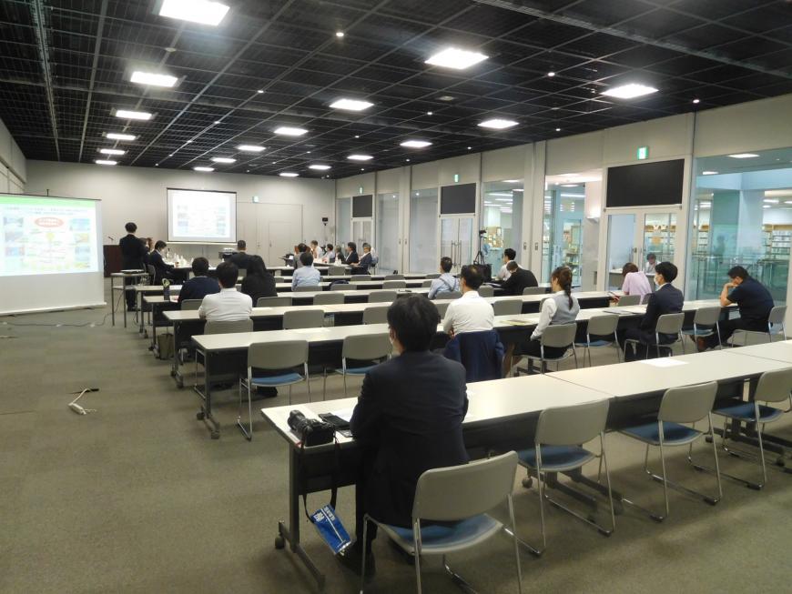 モノクロの室内で参加者の人たちが長机に座り、前方と中央にあるモニターを見ながら説明を聞いている様子を後方から写した写真