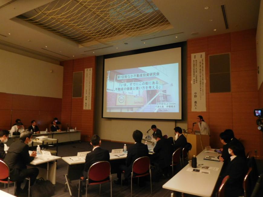 前方に大きなスクリーンがあり右前方の演台に女性が立ちながら身を乗り出し説明をしている様子を写した写真