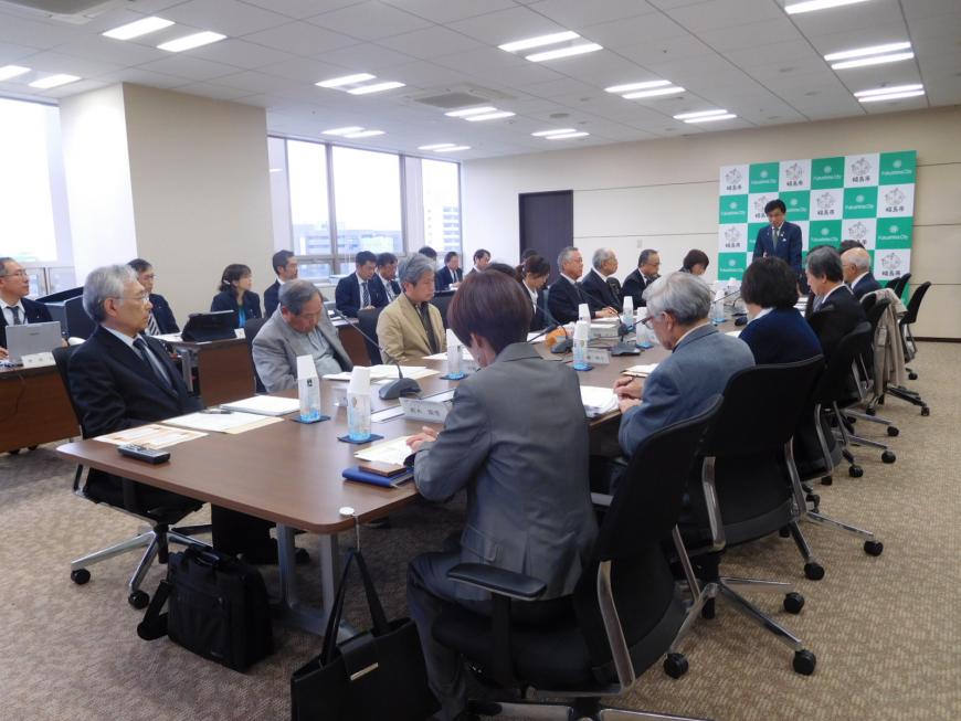 市長がバックボードの前に立ち、向かい合わせに設置された机に座ったたくさんの参加者の人たちが説明を聞いている写真