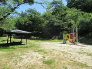 緑に生い茂った木々に囲まれた複合遊具と、傍に休憩所が設置されている公園内の写真