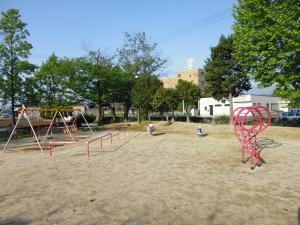 周りを木で囲まれたブランコとスプリング遊具、そしてピンク色の遊具が設置されている公園内の写真