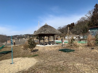 駐車場の隣に茅葺屋の休憩所とパークゴルフ場を囲むように遊歩道が整備されている写真