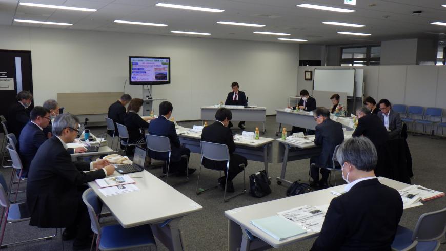 半円状に組み合わせたテーブルの前に座っている関係者とその周りにL字型にテーブルが設置されその前に関係者数名が着席している会議の様子の写真