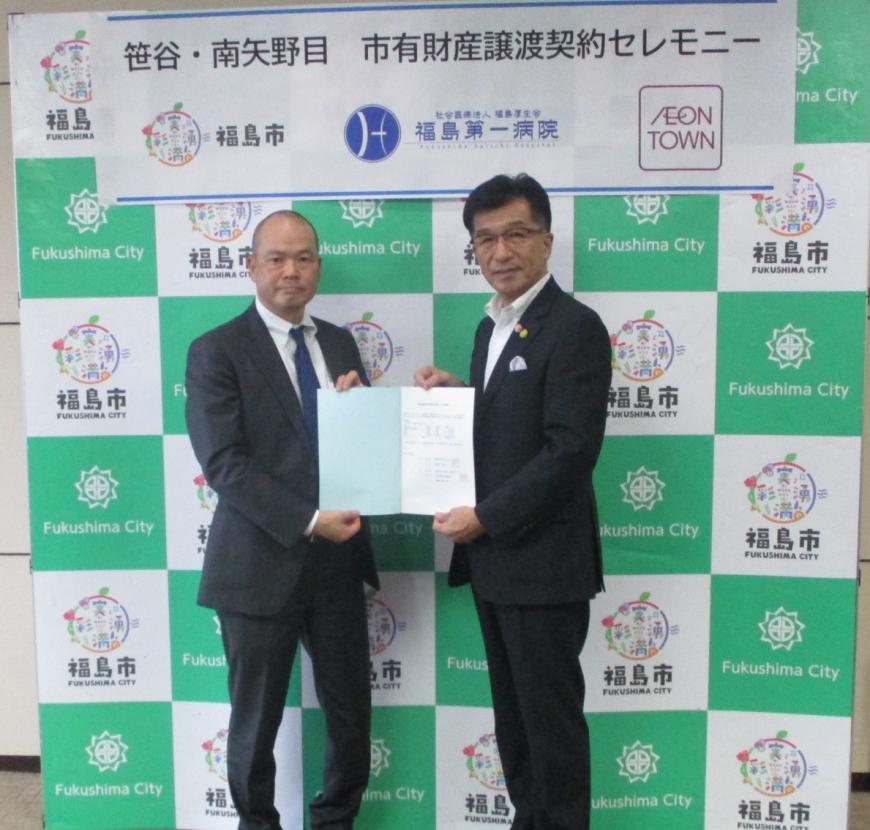 福島市のバックパネルの前に立って契約書を一緒にもって立っている星野俊吾様と木幡市長の写真