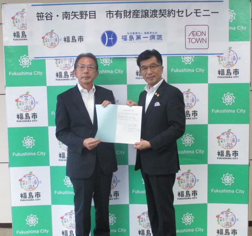 福島市のバックパネルの前に立って契約書を一緒にもって立っている加藤久誠様と木幡市長の写真