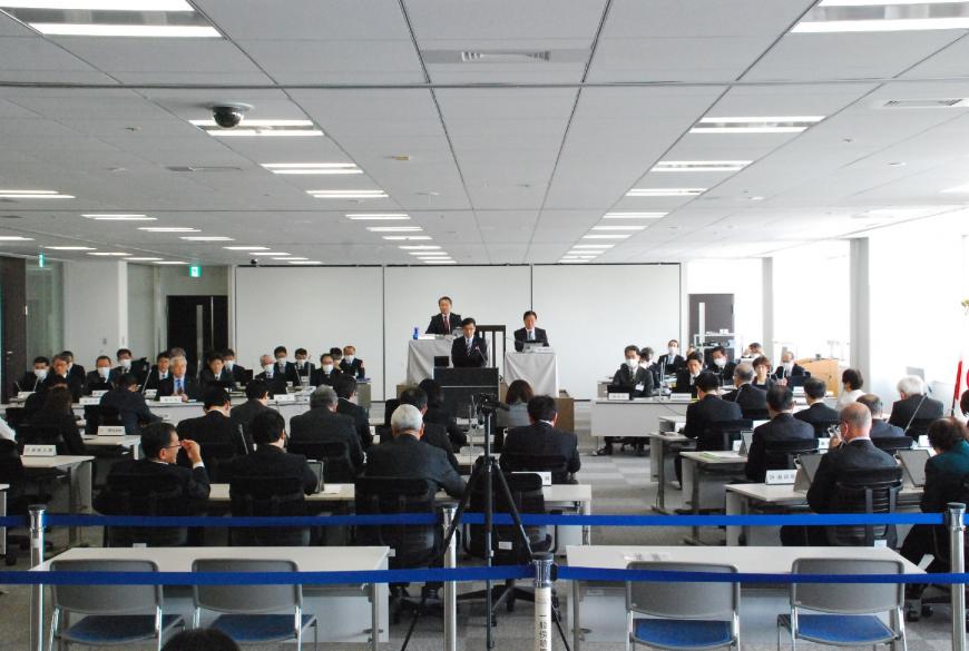 広い会議室で、50名ほどの関係者が席に座り、壇上に設置された卓上マイクで話している写真