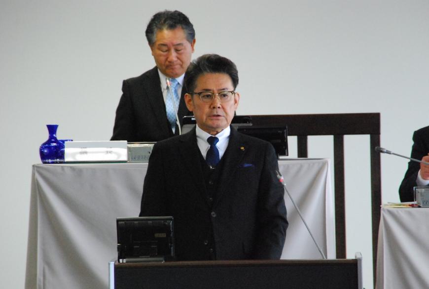 前方の壇上の卓上マイクに向かい話をしている小松良行議員の写真