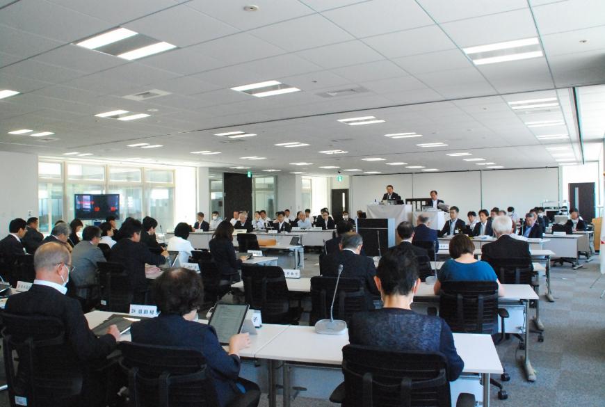 室内中央あたりに設置された演台、並べられた長机の席に多くの関係者の方が座り、6月定例会議が行われている様子を室内後方から写した写真
