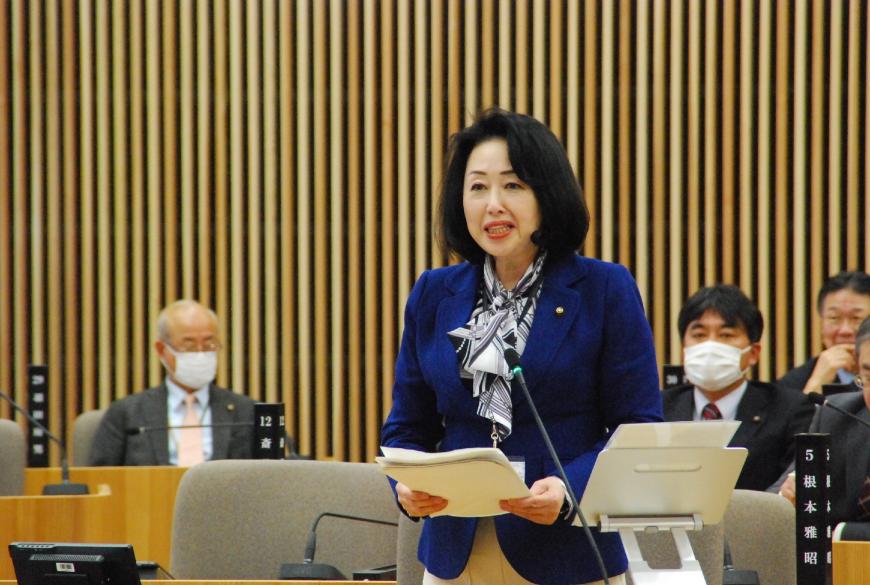 青いジャケットを着た菅原美智子議員が資料を両手で持ち真っすぐ前を向いて話をしている写真