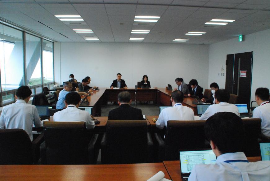 会議室でコの字型に配置された机と横2列に並んだ机の席に座った出席者たちが、各自ノートパソコンの画面を見ながら協議をしている写真