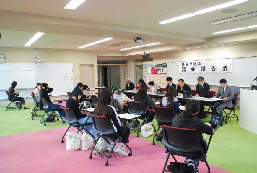 ホワイトボードに「議会報告会」と書かれた横断幕が貼られた部屋で長机に座った議会メンバー7人と、向かい合って席に座った女性団体の参加者たちの写真