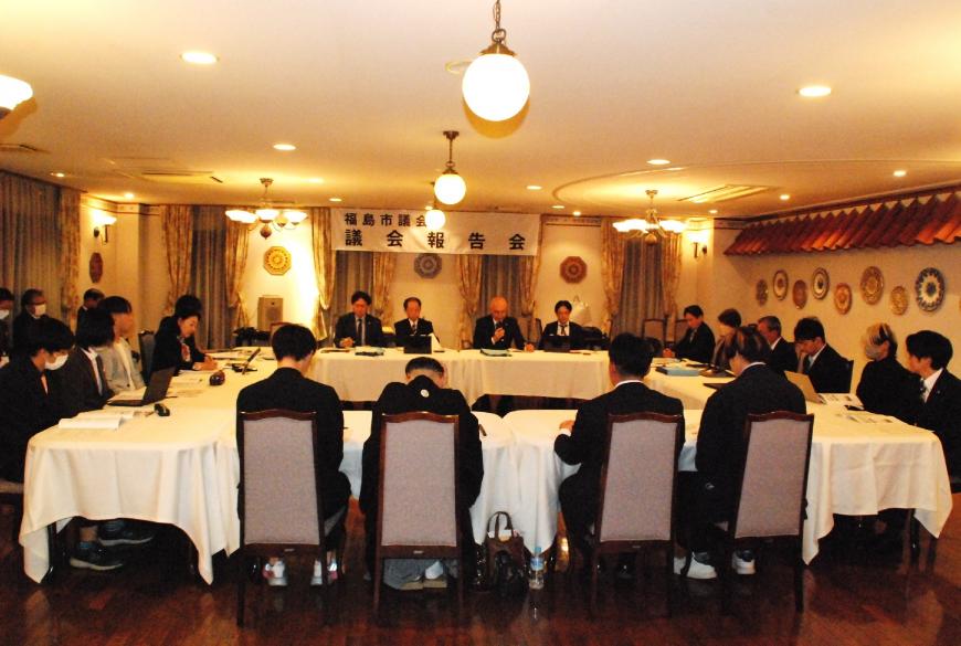 「議会報告会」と書かれた横断幕が掲げられた部屋で、口の字型に配置された白いテーブルクロスが掛けられた席に参加者と議会メンバーが座っている報告会・意見交換会の写真