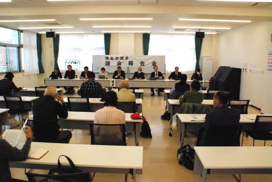「議会報告会」の横断幕が設置された部屋で、数列並んだ長机の席に参加者たちが座り、対面に設けられた席に議会メンバーが座っている写真