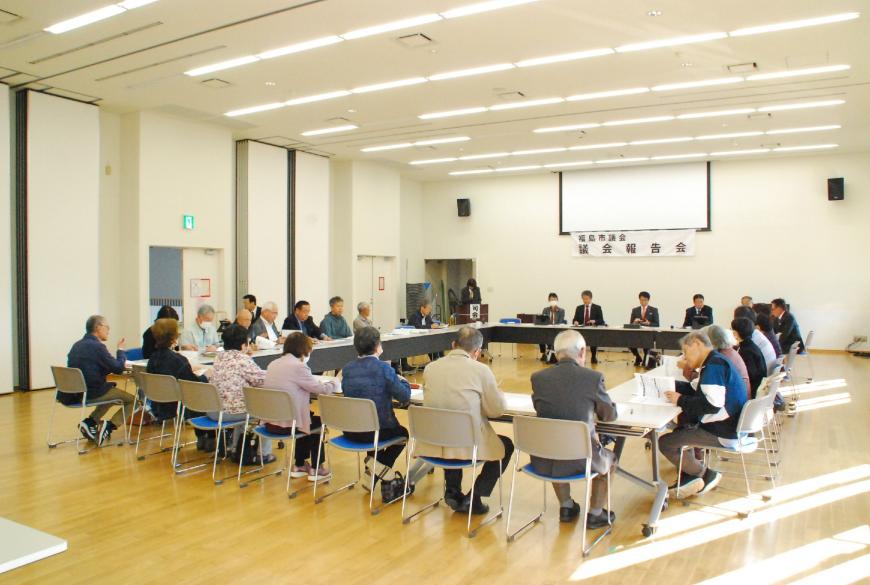 壁に「議会報告会」と書かれた横断幕が掲げられた広い室内に口の字型に配置された机に議会メンバーとたくさんの参加者が座っている写真