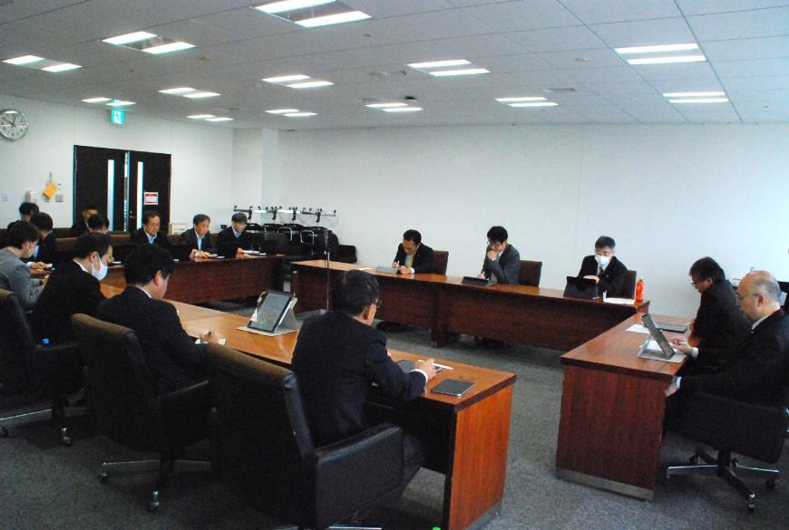 ロの字型に配置された机に女性1人、男性13人の委員が座り、タブレットの画面を見ている写真