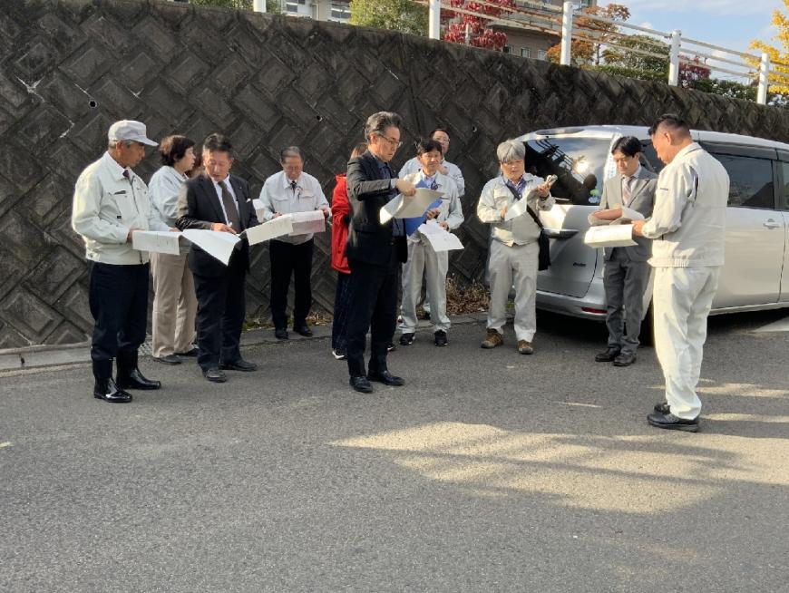 屋外の現場でスーツ姿の男性3人と作業服などを着た男女8人がそれぞれ手に持った資料を見ている写真