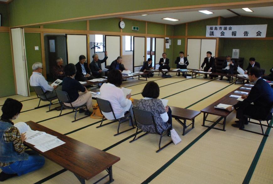 和室の部屋に長机と椅子が設置され参加者が座り、一人の男性が資料を手に持ち、もう片方の手を上げ質問をしている様子の写真