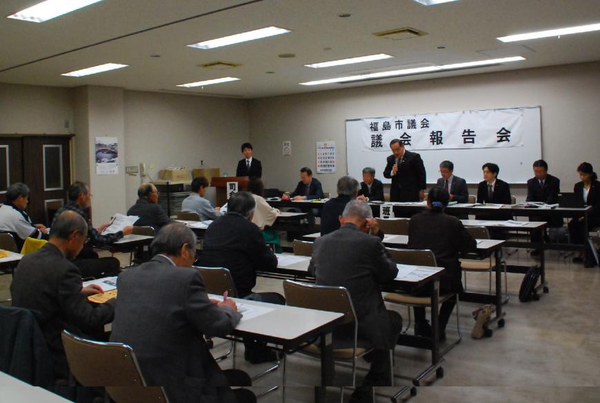 室内に配置されたテーブルと椅子に座り、前方の男性が立ちマイクを使って話をしている様子の写真