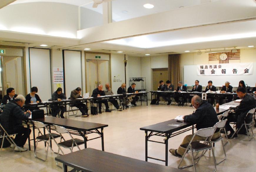 前方の白板に福島市議会議会報告会と掲示され、関係者の方々と参加者の方々がロの字に設置された机の席に着き、資料に目を通しながら会議が行われている様子の写真