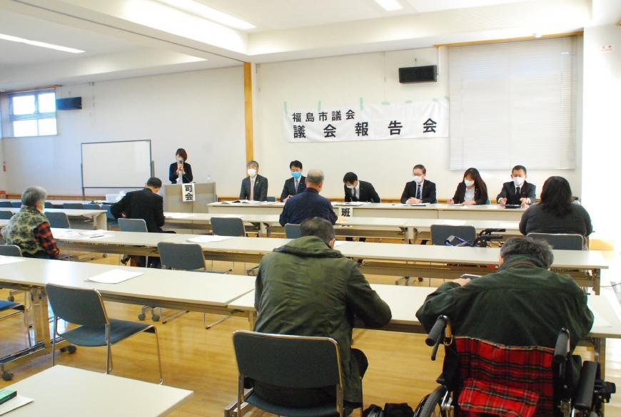 「福島市議会 議会報告会」と書かれた横断幕の前に議員席が設置され、その横の演台に立っている司会の女性がマイクを使い話す様子を議会席の前の座席に参加者が座り聞いている写真