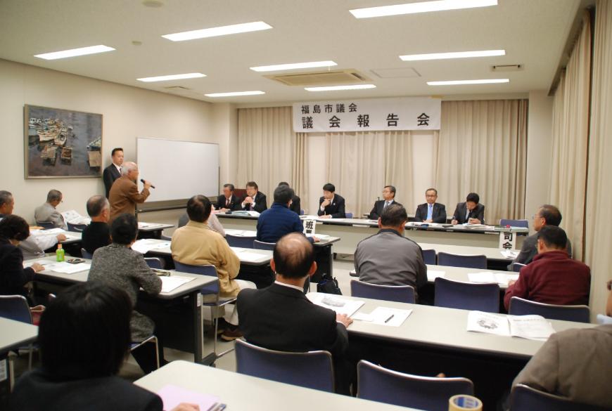 前列に座る司会者や班長の方を向き、参加者の男性が立ってマイクを持ち話をしている様子の写真