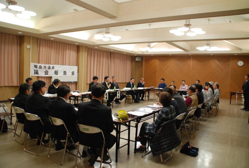 室内にロの字型に設置されたテーブルと椅子に参加者が座り、司会者の方が話す様子を聞いている写真