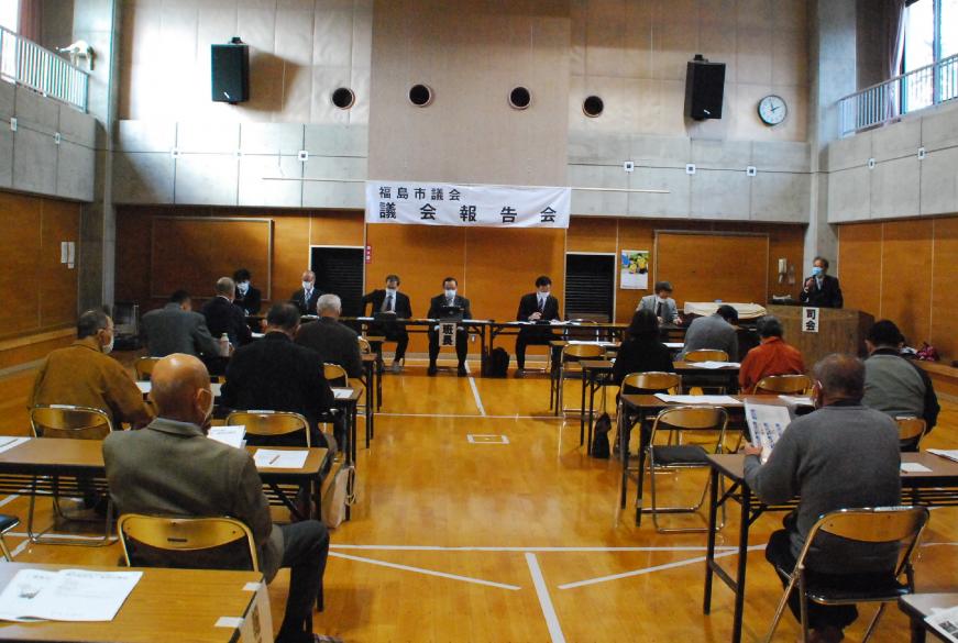 福島市議会議会報告会と書かれた横断幕が前方に掲示され、関係者の方々が前方の長机の前に座り、参加者の方々が会場内の席に着き、意見交換会が行われている様子の写真
