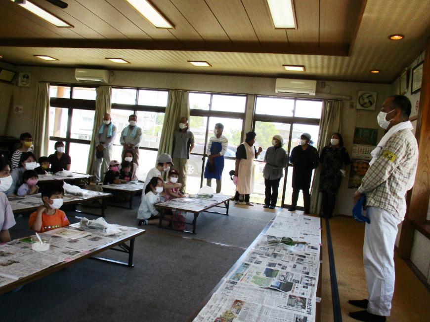 新聞紙が敷かれた長机の後ろに座った子供たちが前方で立って話をする男性の方を見ている写真