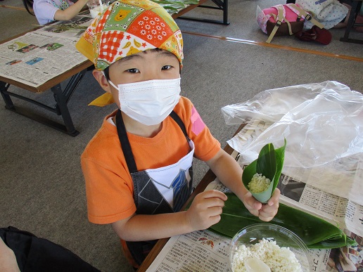 三角巾とマスク、エプロンを身に着けた男の子がもち米が入った笹の葉の筒を手に持っている写真
