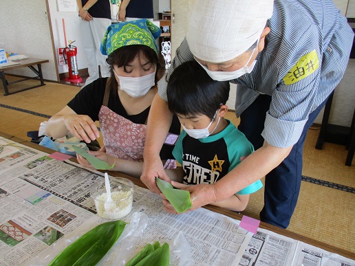 三角巾とマスク、エプロンを身に着けた女性が見守る隣で笹の葉を折りたたむ男の子を後ろから女性がサポートしている様子の写真