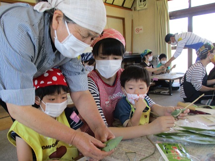 三角巾とマスク、エプロンを身に着けた家族が見つめる中、女性が子どもの頭越しに結び方を教えている写真