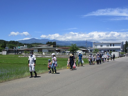 青空の下、田んぼ横の道路を一列になって歩く参加者たちの写真