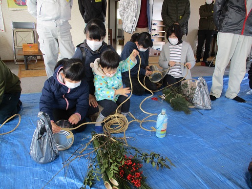女性の膝に座った男の子が伸ばした縄を、隣に座った女の子が缶に巻き付けている様子の写真
