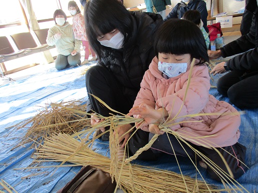 小さな女の子が両手で持ったわらの束をねじり、それを後ろの女性が見守っている写真