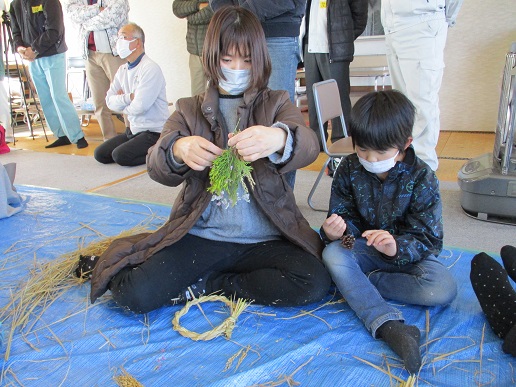 わらでできた輪っかを足元に置き、松ぼっくりや草花を手に持っている親子の写真