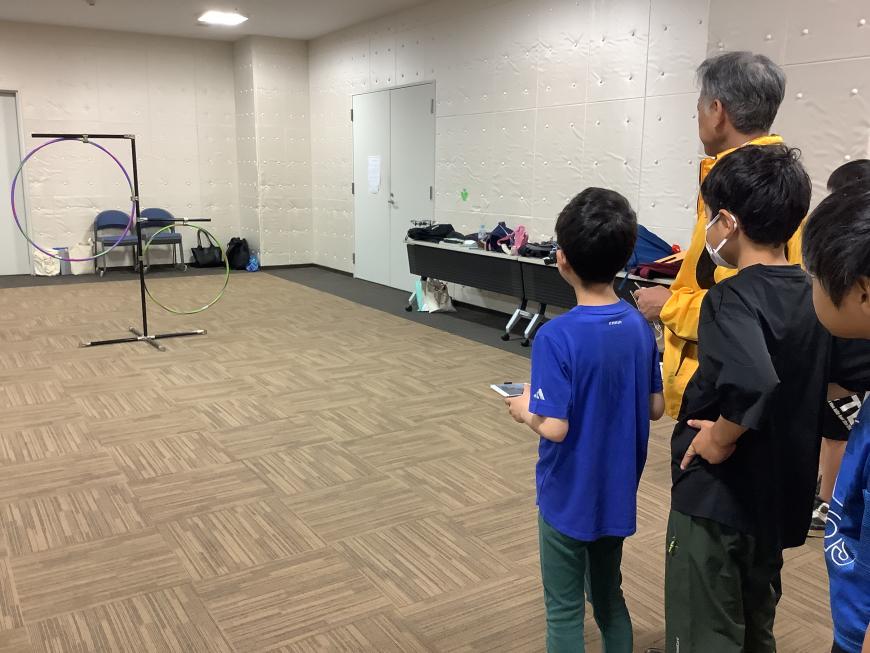 子ども達が整列している少し離れた向かい側に、大きさの異なるフラフープが吊るされている写真