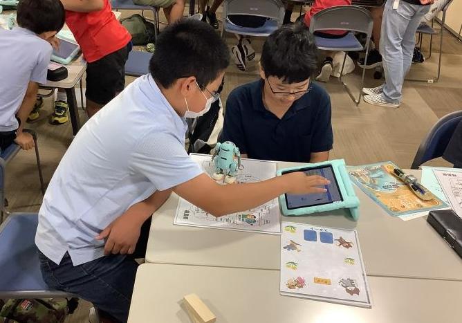 席についている男の子が同じテーブルの席についている男の子が操作しているタブレットを見ている写真