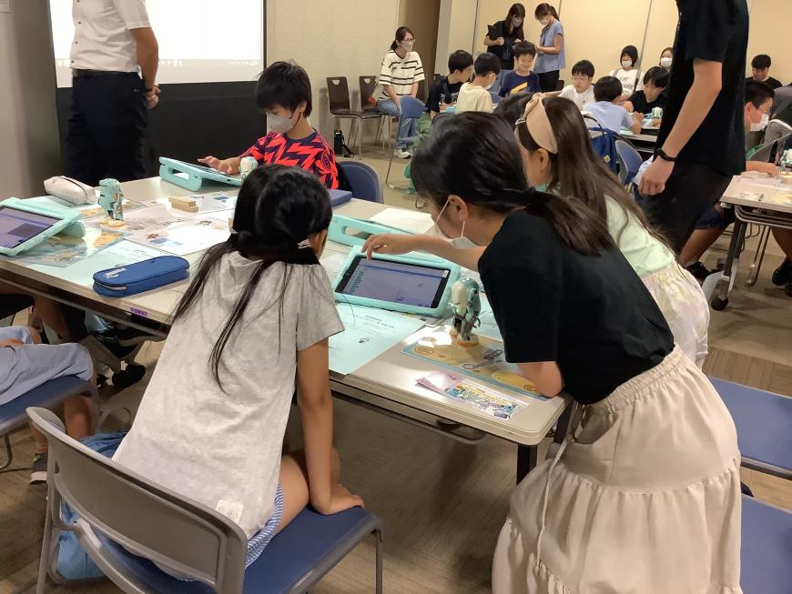 1人の女の子が体を前のめりになってタブレット操作をし、もう一人の女の子がその様子を椅子に座り見ている写真