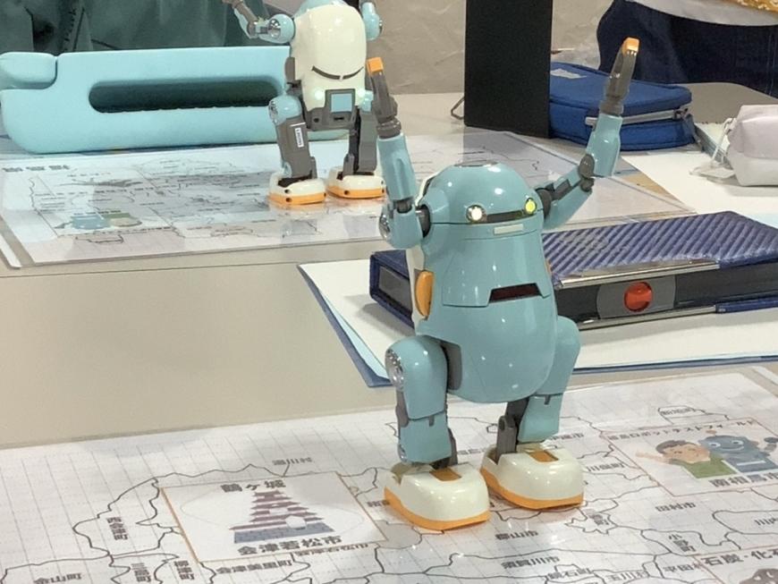 机の上に置かれた2台の二足歩行ロボット「あるくメカトロウィーゴ」が両手をあげている写真