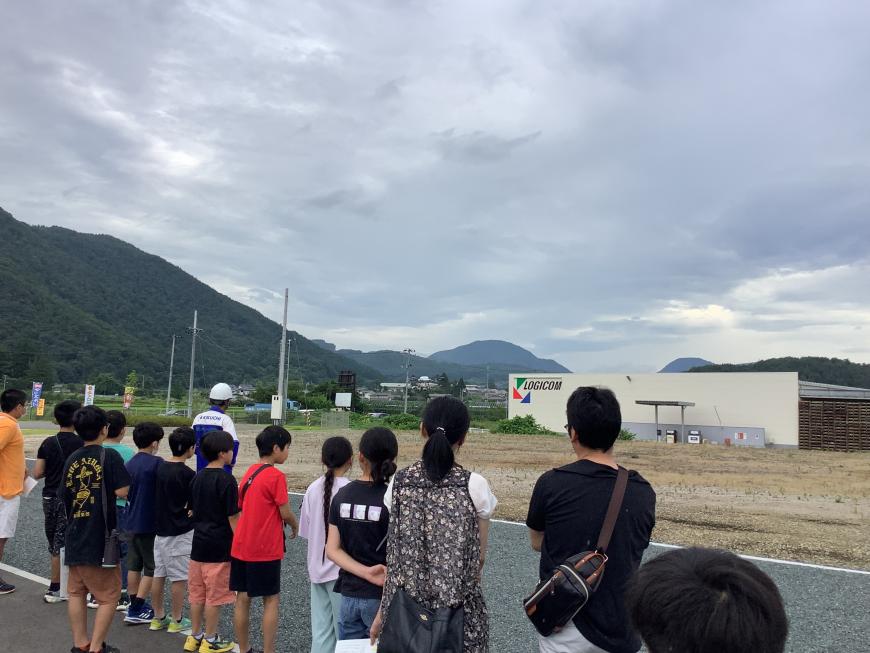 奥に山や、工場のような大きな建物が見える広い敷地に、参加者たちが横一列に並び広い敷地の方を見ている写真