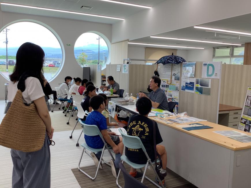 体験ブースに設けられたパイプ椅子に座っている子供たちが、体験ブースにいる男性の話を聞いている様子の写真