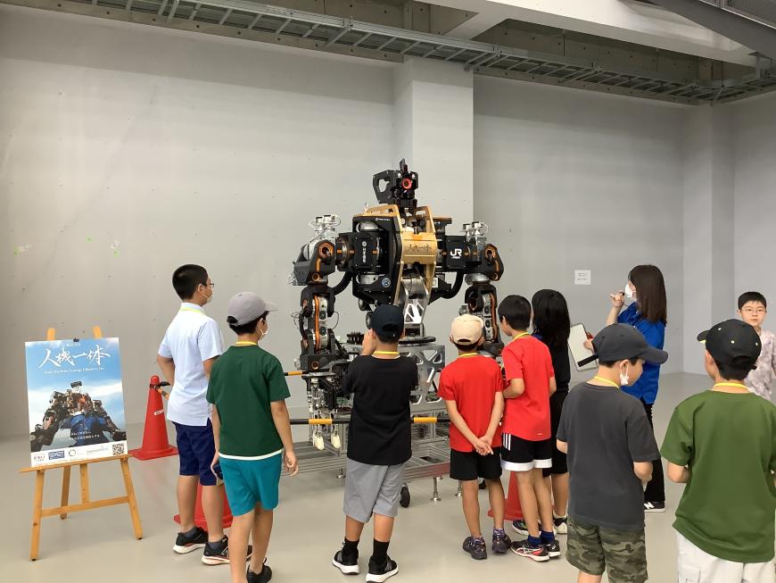 ロボットの写真と「人機一体」と書かれたポスターの横に展示されている大きなロボットを見学している子供たちの写真
